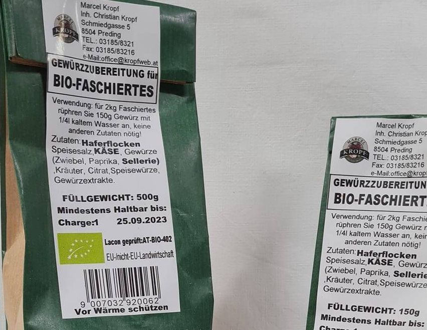 Zwei Beutel Bio-Gewürzmischung, einer geöffnet und einer geschlossen, liegen auf einem Tisch. Daneben befinden sich ein Teller mit Gewürzmischung und ein Teller mit geschnittenem Fleisch.