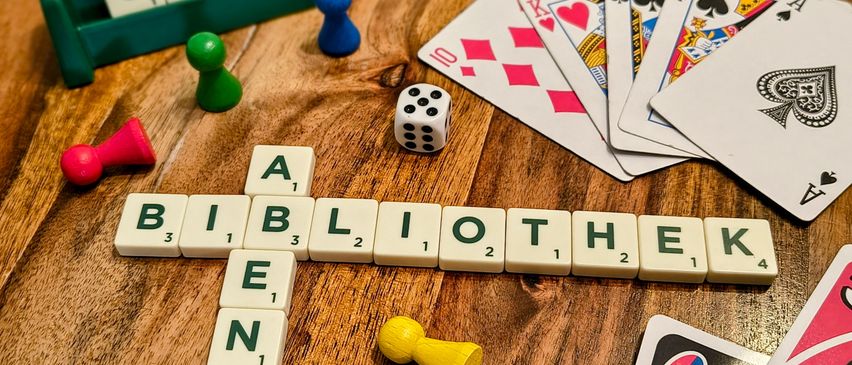 Ein Holztisch mit einem Scrabble-Spiel, dem Wort 'Bibliotheke', Würfeln, Spielsteinen und Spielkarten.