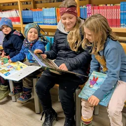 Eine Gruppe von Kindern und einer Frau sitzen auf Stühlen in einer Bibliothek und lesen Bücher. Die Frau liest den Kindern ein Buch vor.