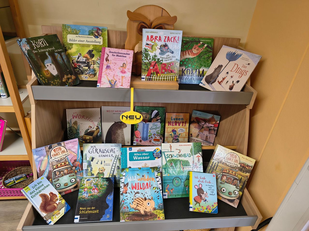 A wooden bookshelf filled with children's books, some with a yellow tag reading 'NEW'. The books have various illustrations, including animals, people, and fantasy themes. The books are arranged in rows on the shelves.