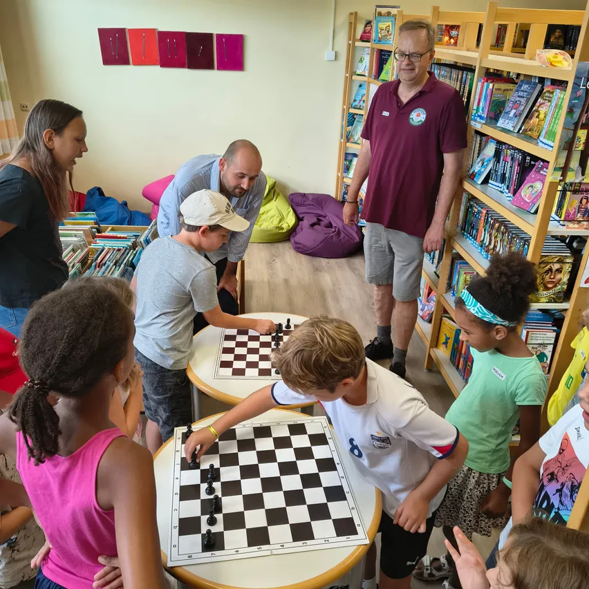 Eine Gruppe von Kindern und Erwachsenen spielt Schach in einer Bibliothek. Zwei Jungen spielen gegeneinander.