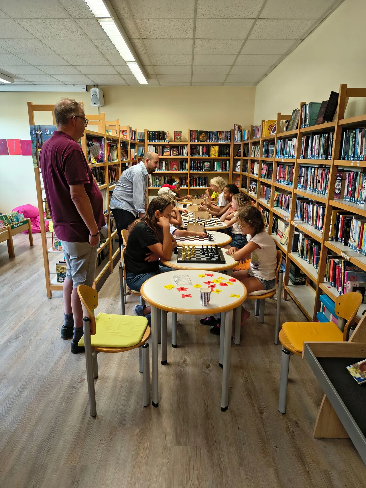 Eine Gruppe von Kindern und Erwachsenen spielt Schach in einer Bibliothek. Die Schachfiguren stehen auf den Tischen, und die Spieler konzentrieren sich auf ihr Spiel.