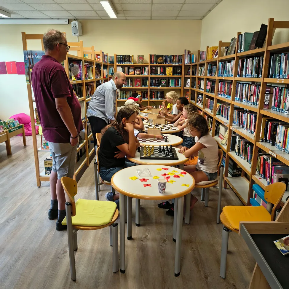 Eine Gruppe von Kindern und Erwachsenen spielt Schach in einer Bibliothek. Die Schachfiguren stehen auf den Tischen, und die Spieler konzentrieren sich auf ihr Spiel.