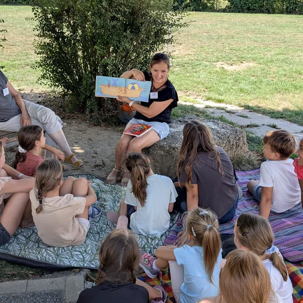 Eine Frau liest ein Buch vor einer Gruppe von Kindern, die in einem Kreis auf einer Decke im Park sitzen.