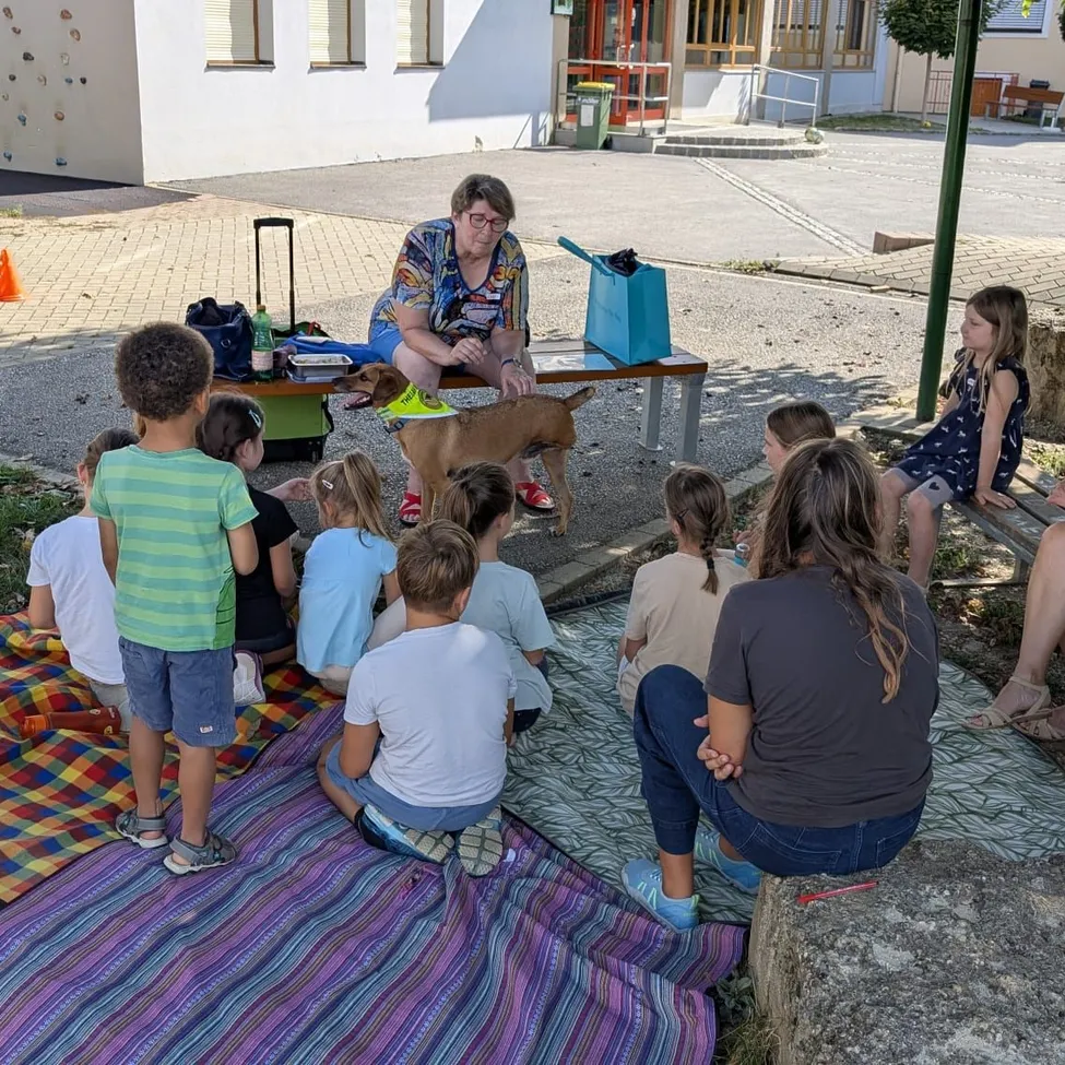 Eine Frau mit Brille sitzt auf einer Bank mit einem Hund, umgeben von Kindern, die auf einer Decke auf dem Boden sitzen.