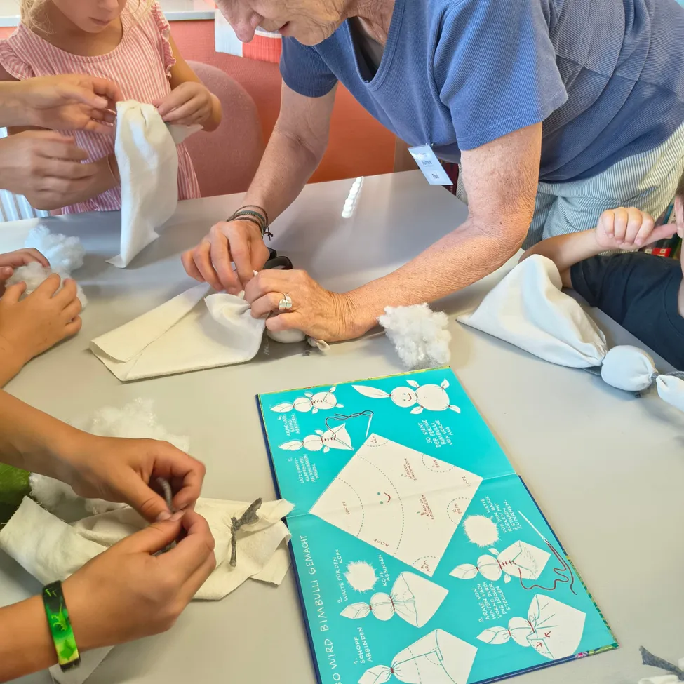 Eine ältere Frau lehrt Kinder das Nähen mit einem blauen Buch mit einer Hasenillustration als Anleitung. Die Kinder sitzen um einen Tisch mit verschiedenen Materialien.