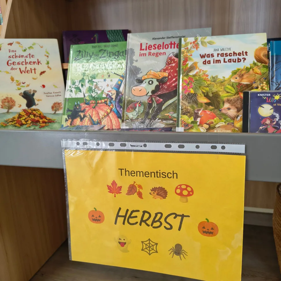 Ein Bücherregal mit verschiedenen Kinderbüchern über Herbst und Halloween. Die Bücher sind in einer Reihe angeordnet, und ein gelbes Schild mit dem Wort 'Herbst' hängt davor.