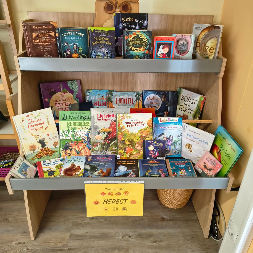 Ein hölzerne Bücherregal voller Kinderbücher, darunter Titel wie 'Unheimliche Geschichten' und 'Halloween Spaß', mit einem gelben Schild, das 'Herbst' besagt.
