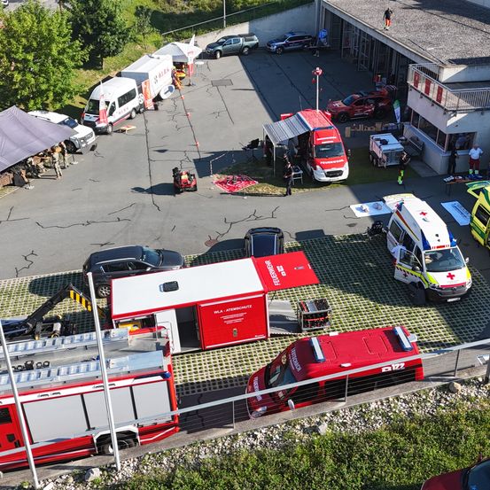 Ein Luftbild eines Notfallbereichs mit mehreren Fahrzeugen, darunter Krankenwagen und Feuerwehrfahrzeuge, die auf einem Parkplatz geparkt sind. Es stehen und gehen Leute herum. Links ist ein Zelt aufgebaut.