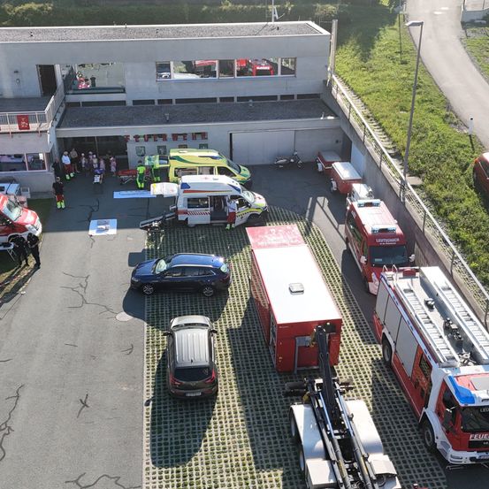 Ein Luftbild einer Feuerwehrwache mit mehreren geparkten Rettungsfahrzeugen, darunter Krankenwagen und Feuerwehrfahrzeuge. Menschen sind in der Nähe des Eingangs versammelt, und das Gebiet ist gut gepflegt mit viel Grün.