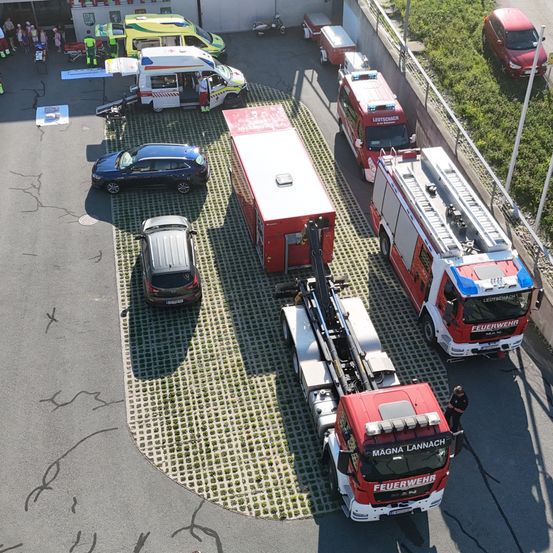 Eine Draufsicht auf Rettungsfahrzeuge in einem Parkplatz, einschließlich Feuerwehrwagen, Krankenwagen und eines Versorgungsfahrzeugs, mit Menschen in der Nähe.