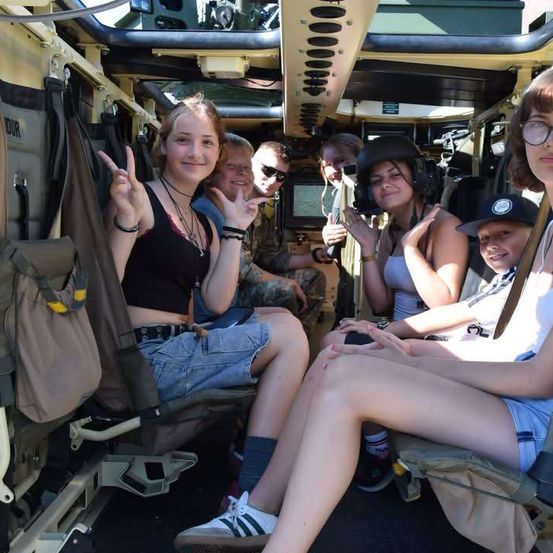 Eine Gruppe von Menschen sitzt in einem Militärfahrzeug und lächelt für ein Foto. Das Fahrzeug hat ein beiges Interieur mit verschiedenen Fächern und Ausrüstungsgegenständen.