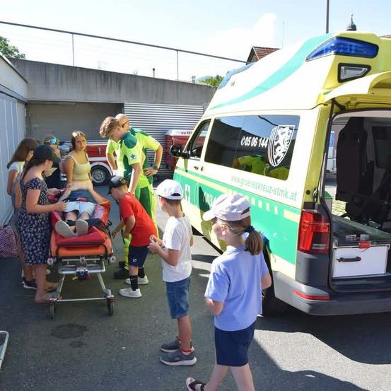 Eine Gruppe von Kindern und Erwachsenen versammelt sich um einen Krankenwagen mit offener Hecktür. Eine Person auf einer Trage wird von medizinischem Personal betreut.