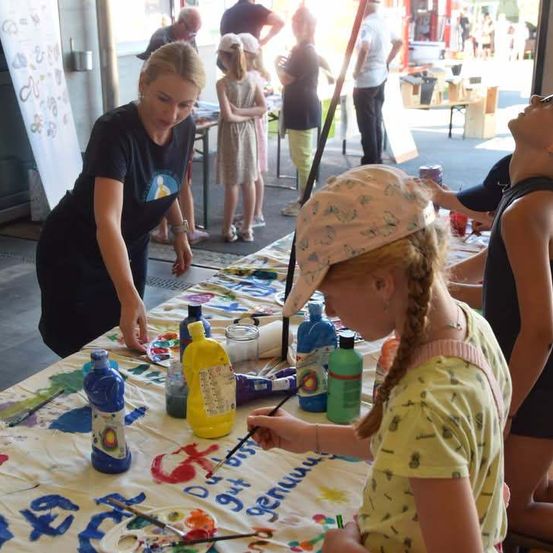Eine Frau hilft einem jungen Mädchen, auf einer großen Leinwand zu malen, bei einer Veranstaltung im Freien. Andere Kinder und Erwachsene sind anwesend, einige stehen und beobachten.