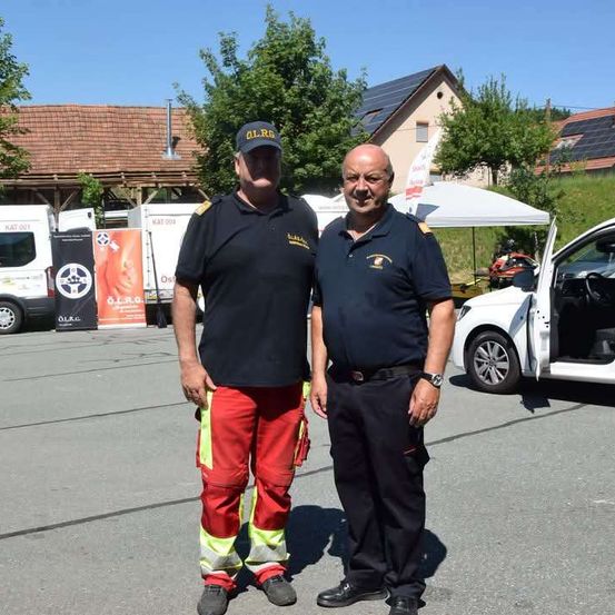 Zwei Männer in Uniform stehen auf einem Parkplatz, einer trägt rote und gelbe Hosen, der andere schwarz. Hinter ihnen ist ein weißes Zelt und ein weißer Van zu sehen. Im Hintergrund befinden sich Häuser mit Solarmodulen auf den Dächern.