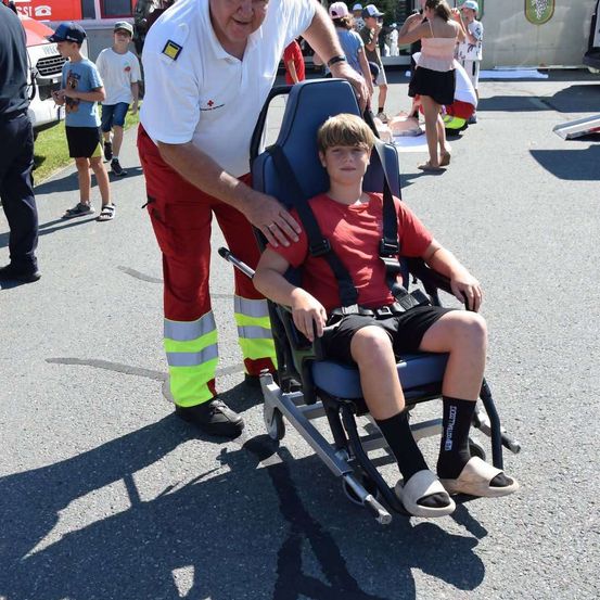 Ein Mann in einer rot-weißen Uniform hilft einem Jungen in einem roten T-Shirt, sich an einem sonnigen Tag in einen Stuhl zu setzen.