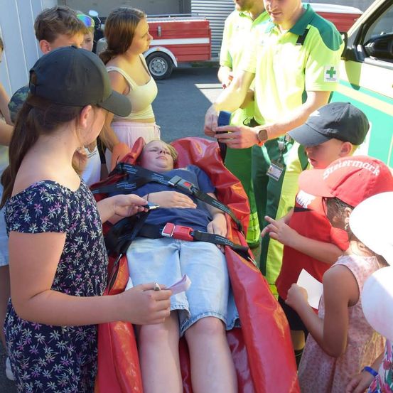 Ein junger Junge liegt auf einer roten Trage, die mit einem Sicherheitsgurt befestigt ist, während mehrere Kinder und Erwachsene, einschließlich eines Mannes in einem grünen Uniform, um ihn herum stehen.