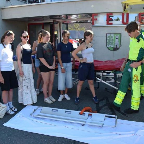 Eine Gruppe junger Frauen steht vor einer Feuerwache und beobachtet einen Sanitäter, der eine Trage demonstriert.