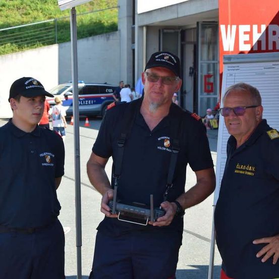 Drei Männer in Uniform stehen draußen, einer hält ein Gerät. Ein Polizeifahrzeug und ein Gebäude mit dem Wort Wehr sind im Hintergrund.
