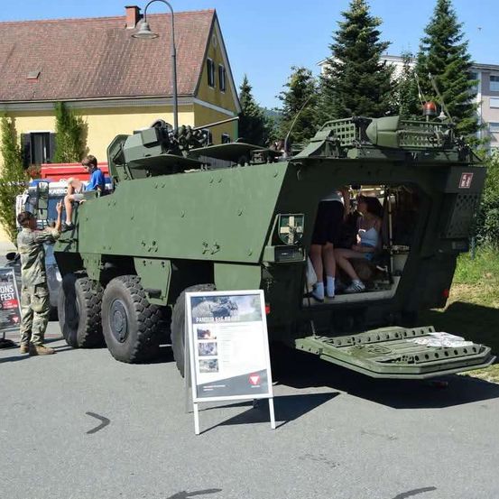 Ein grünes Militärfahrzeug mit montierter Kanone ist am Straßenrand geparkt. Ein Soldat steht in der Nähe, und Menschen sitzen im Inneren des Fahrzeugs. Ein Schild steht vor dem Fahrzeug.