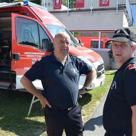 Zwei Feuerwehrleute stehen vor einem roten Krankenwagen, einer trägt eine Uhr und der andere eine Kappe. Hinter ihnen ist ein Gebäude mit Balkon und einer roten Fahne zu sehen.