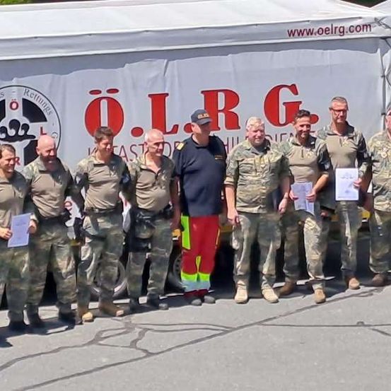 Eine Gruppe von Personen in Camouflage-Uniformen steht vor einem weißen Zelt mit dem Logo 'O.L.R.G.' und einer Website-Adresse. Sie halten Urkunden in den Händen und scheinen für ein Foto zu posieren.