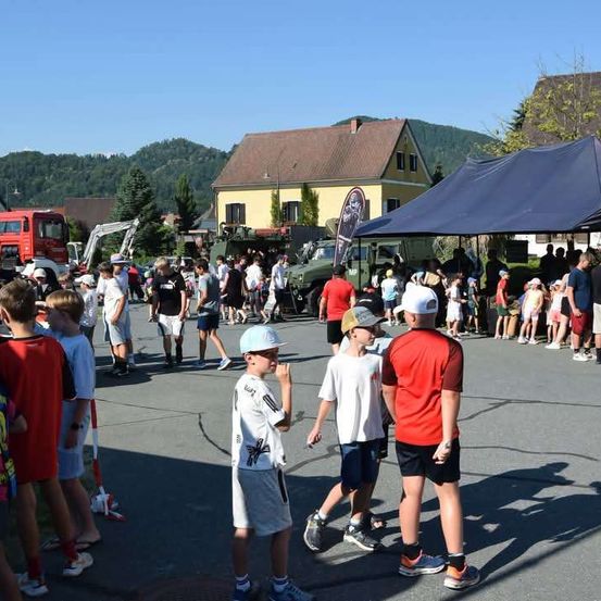 Eine Gruppe von Kindern und Erwachsenen versammelt sich in einem Außenbereich, möglicherweise für eine Veranstaltung. Einige tragen Hüte und Turnschuhe. In der Nähe sind einige Militärfahrzeuge geparkt.