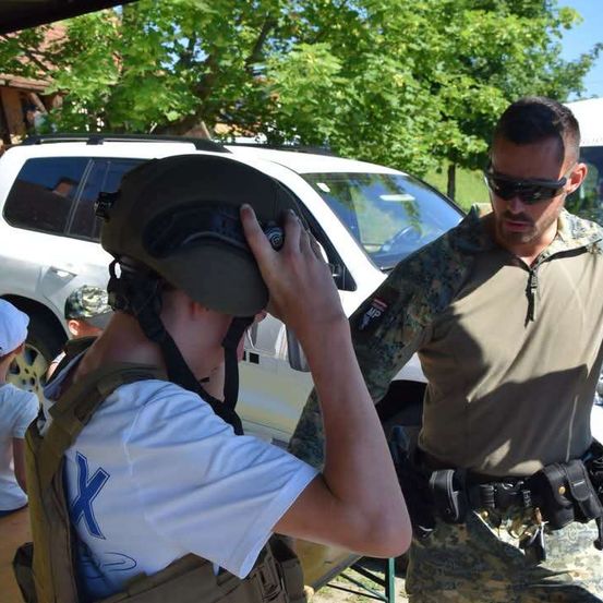 Ein Mann in Uniform hilft einem Jungen, einen Helm aufzusetzen. Sie stehen neben einem weißen SUV.