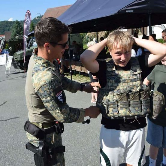 Ein Soldat in Tarnuniform hilft einem jungen Jungen, eine kugelsichere Weste in einer Außenumgebung anzulegen.