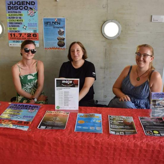 Drei Frauen sitzen an einem Tisch mit Broschüren und lächeln für ein Foto. Hinter ihnen werben Poster für Veranstaltungen und eine Disco.