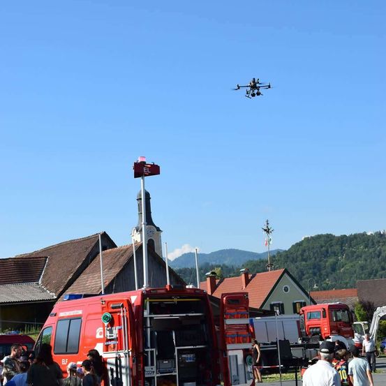 Ein rotes Notfallfahrzeug steht in einem Dorf, über dem ein Drohnen fliegt. Mehrere Menschen sind versammelt, und es gibt andere Fahrzeuge und Gebäude im Hintergrund.