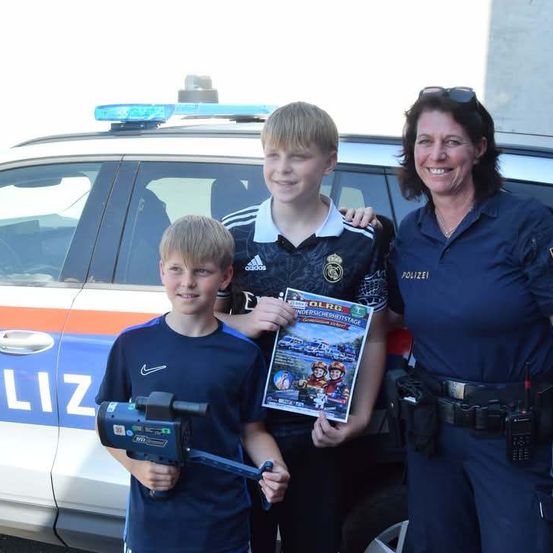 Zwei Jungen und eine Polizistin stehen vor einem Polizeiauto, ein Junge hält eine Kamera und eine Zeitschrift, der andere eine Zeitschrift, und die Polizistin lächelt.