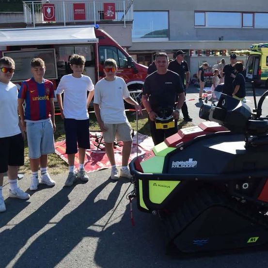 Vier Jungen und ein Mann stehen neben einem roten Fahrzeug, möglicherweise einem Rettungsfahrzeug, mit einem Krankenwagen und einem Gebäude im Hintergrund.