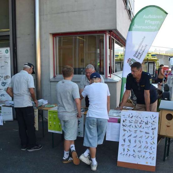 Eine Gruppe von Menschen steht um einen Tisch mit Vogelhäusern und Postern herum, möglicherweise bei einer Gemeinschaftsveranstaltung. Eine Person schaut sich einen Plakat an, während andere in ein Gespräch vertieft sind.