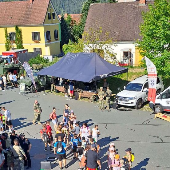 Eine Menschenmenge, darunter Kinder, versammelt sich um ein Militärzelt und Fahrzeuge auf einem Stadtplatz. Einige tragen Rucksäcke und Hüte.
