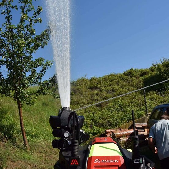 Ein Wassersprüher sprüht Wasser aus einer rot-schwarzen Maschine auf einen Baum in einem Feld unter einem klaren blauen Himmel.