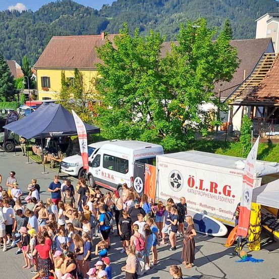 Eine Menschenmenge versammelt sich um zwei weiße Trucks, einer mit dem Text 'O.L.R.G.' und der andere mit 'Katastrophenhilfedienst', in einem Dorf mit Häusern und Bergen im Hintergrund.
