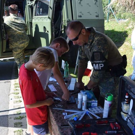 Zwei Jungen arbeiten an einem Tisch mit verschiedenen Werkzeugen und Gegenständen, während ein Militärpolizist sie beobachtet. Im Hintergrund ist ein Militärfahrzeug geparkt.