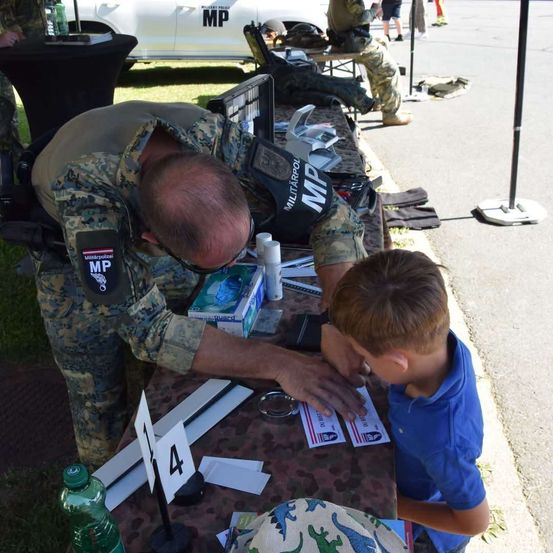 Ein Militärpolizist zeigt einem kleinen Jungen ein Dokument auf einem Tisch. Der Polizist trägt eine Camouflage-Uniform und der Junge trägt ein blaues Hemd.
