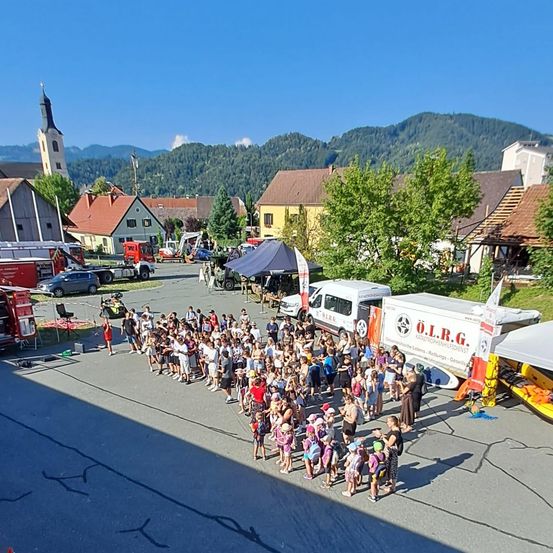 Eine große Gruppe von Menschen, einschließlich Kindern, versammelt sich auf einem Stadtplatz mit Bergen im Hintergrund. Verschiedene Fahrzeuge und Zelte sind vorhanden.