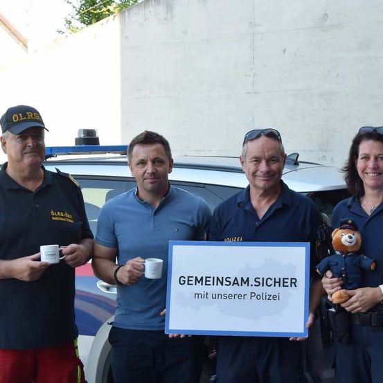 Vier Personen, zwei Männer und zwei Frauen, stehen vor einem Polizeiauto. Sie halten Tassen und lächeln für die Kamera. Ein Schild in der Mitte steht 'GEMEINSAM.SICHER mit unserer Polizei'.