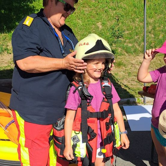 Eine Frau in Uniform hilft einem kleinen Mädchen, einen gelben Helm aufzusetzen. Das Mädchen trägt einen roten Rettungsweste und lächelt. Eine andere Mädchen steht in der Nähe und trägt einen rosa Hut.