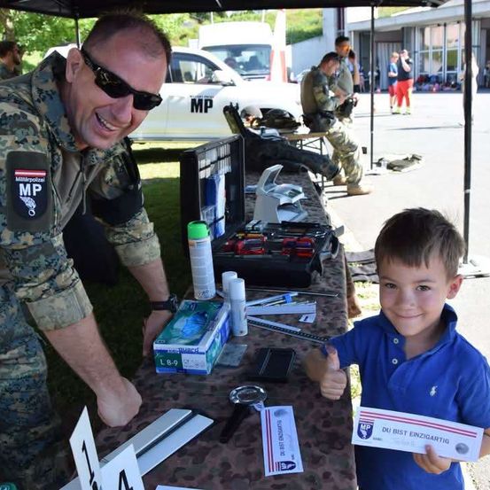Ein Militärpolizist zeigt einem kleinen Jungen eine Urkunde, während er an einem Tisch mit verschiedenen Gegenständen steht.