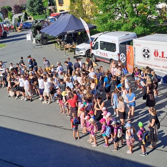 Eine Menschenmenge, wahrscheinlich Schüler, versammelt sich im Freien. Einige tragen Hüte und haben Rucksäcke dabei. Fahrzeuge, darunter ein Van und ein LKW, sind in der Nähe geparkt. Ein Zelt ist aufgebaut, mit einer Person darin.