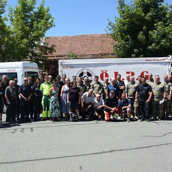 Eine Gruppe von Menschen in Uniformen und Freizeitkleidung posiert für ein Foto vor zwei Fahrzeugen, eines davon mit dem Text 'O.J.R.G.'. Dahinter steht ein Gebäude mit einem roten Dach und Bäumen.