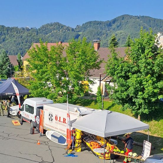Ein weißer Lieferwagen mit den Buchstaben O.L.R. am Straßenrand geparkt, darunter ein Zelt und ein Boot. Eine Person steht unter dem Zelt. Im Hintergrund befindet sich ein Haus mit Schornstein und Bäumen.