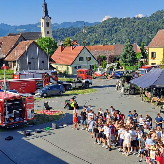Eine Gruppe von Menschen steht in einer Reihe vor einem Dorf mit Bergen im Hintergrund. Fahrzeuge, einschließlich Feuerwehrwagen und Maschinen, sind in der Gegend geparkt.