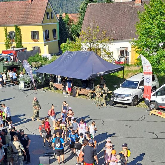 Eine Menschenmenge versammelt sich auf einem Stadtplatz mit aufgestellten Militärfahrzeugen und Zelten. Einige Personen tragen Uniformen, andere sind in Freizeitkleidung. Bäume und Gebäude sind im Hintergrund sichtbar.