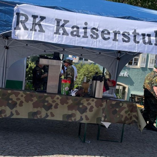 Ein Tisch mit Tarnmuster unter einem Zelt mit einem Banner, das 'RK Kaiserstuhl' steht. Zwei Personen in Militäruniformen stehen hinter dem Tisch, mit verschiedenen Gegenständen darauf. Eine weitere Person in Uniform steht rechts.