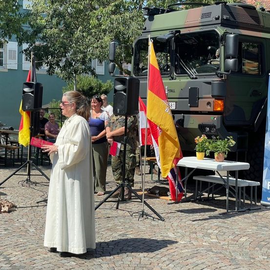 Eine Frau in einem weißen Gewand steht vor einem Militärfahrzeug und hält ein Buch. Hinter ihr sind eine Flagge und ein Lautsprecher aufgestellt. Einige Leute sind versammelt, einige in Uniform.